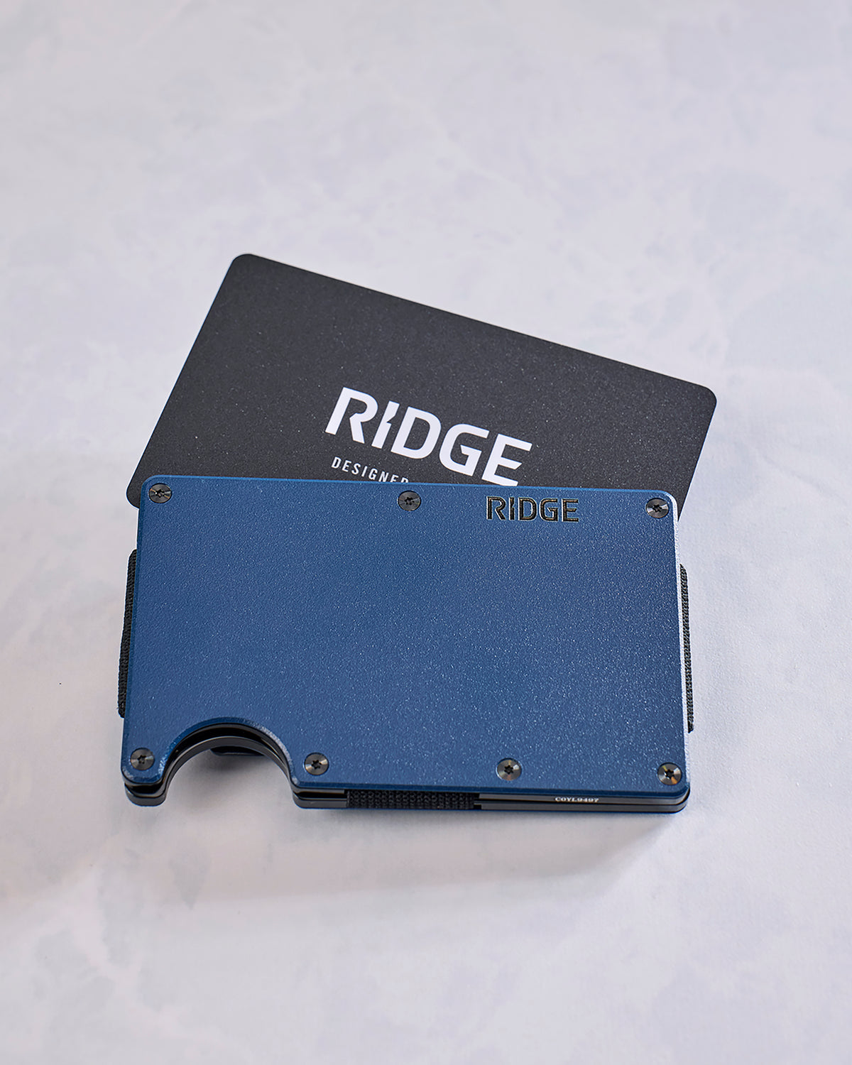 Back view of a navy Ridge wallet with exposed screws, a card extending from the top, and a thumb notch at the bottom.