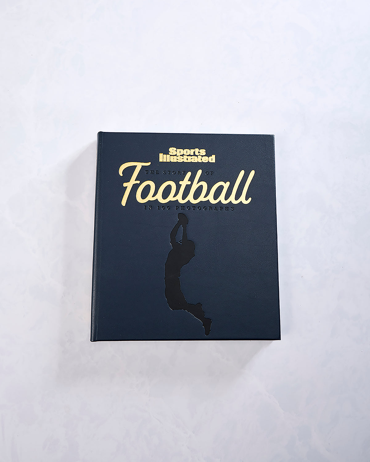 Closed navy book titled “The Story of Football in 100 Photographs” from Sports Illustrated, with a gold silhouette of a football player.