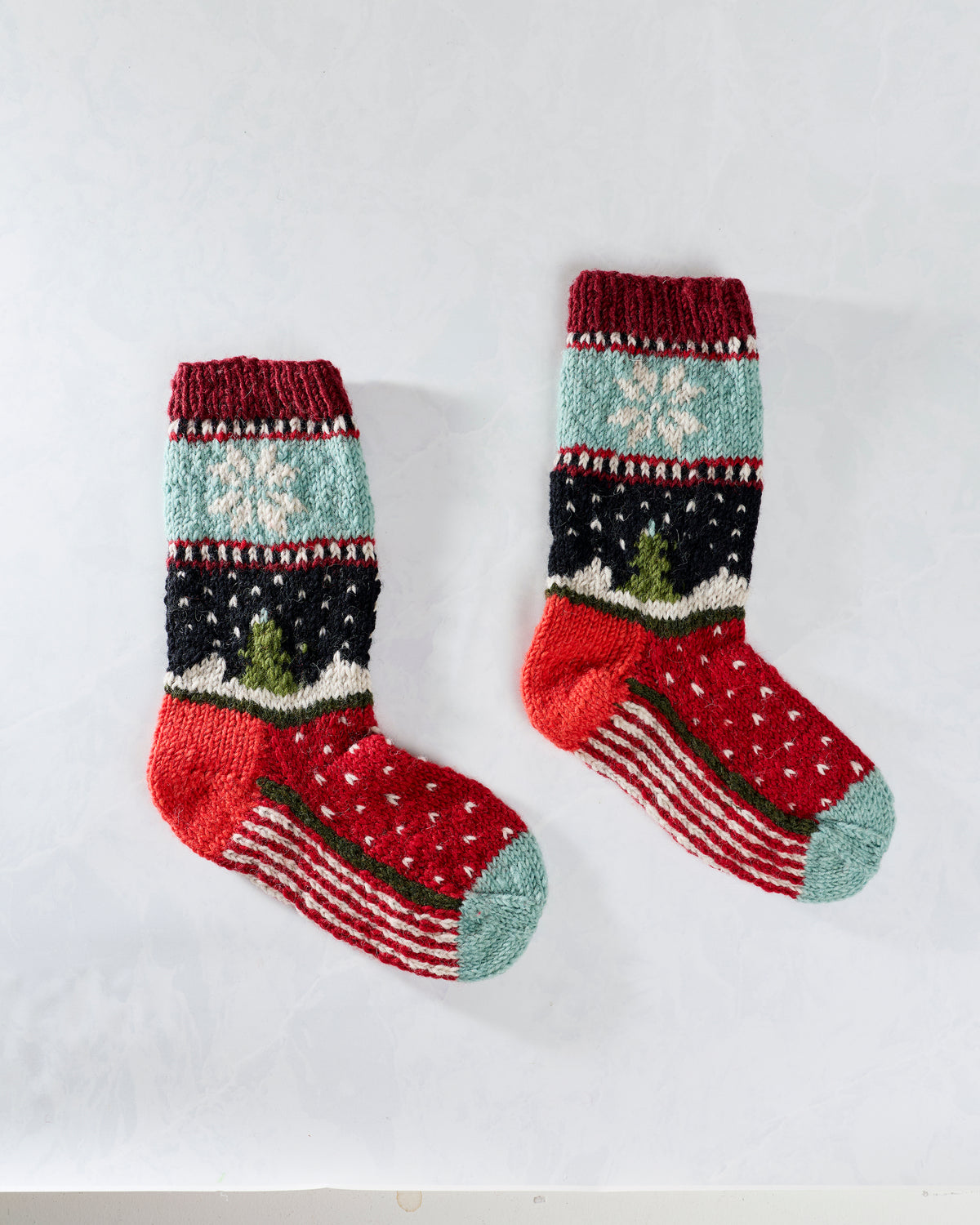 Pair of multicolored knitted holiday socks displayed separately, with snowflake and tree motifs.