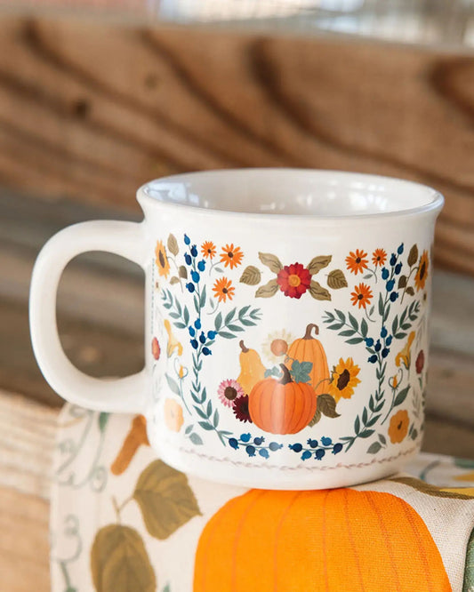 White mug with a colorful floral border and illustrations of pumpkins, flowers, and berries, shown in a fall-themed setting.