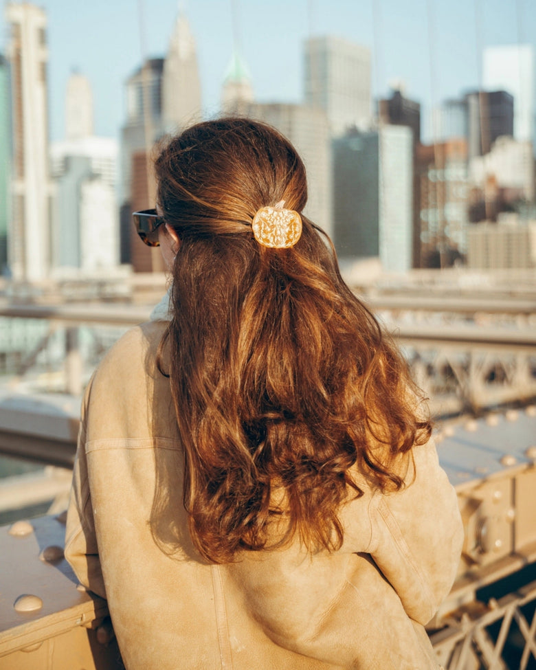 Orange pumpkin-shaped claw hair clip securing a half-up hairstyle, shown from behind against a city skyline.