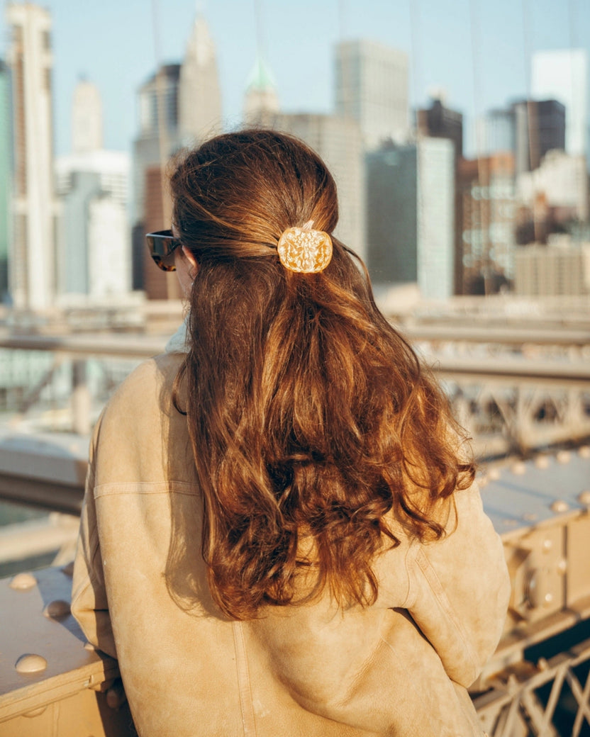 Orange pumpkin-shaped claw hair clip securing a half-up hairstyle, shown from behind against a city skyline.
