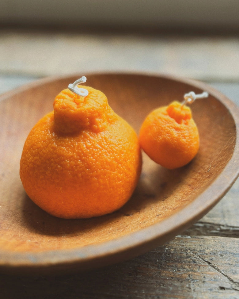 Orange sumo orange beeswax candle beside a smaller orange candle on a wooden plate.