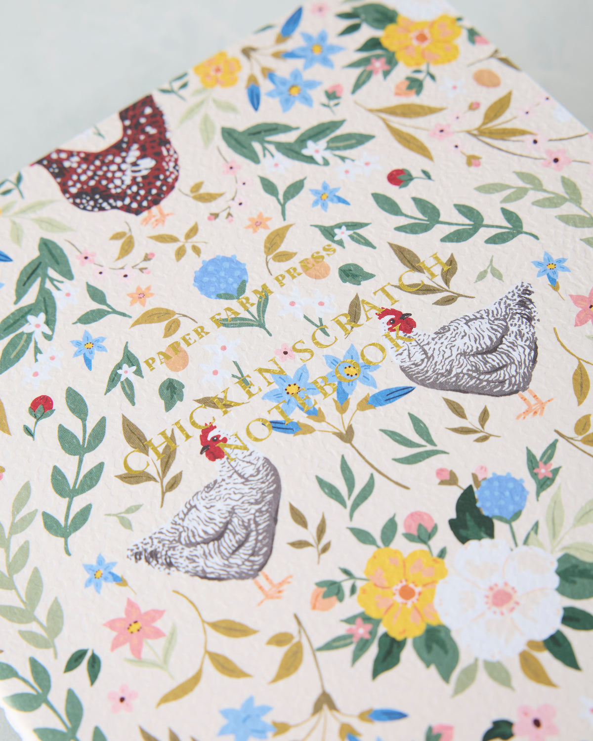 Close-up of the Chickens & Flowers notebook cover with illustrated chickens, flowers, leaves, and gold lettering.