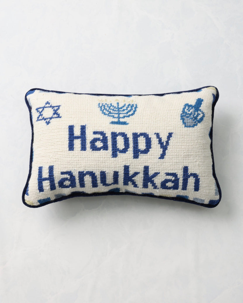 Front of an ivory needlepoint pillow reading “Happy Hanukkah,” with a Star of David, menorah, and dreidel motifs.