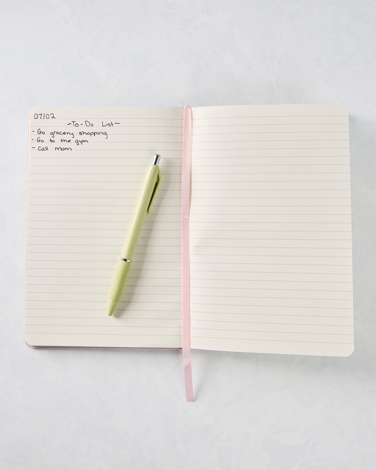 Open lined journal with a pink ribbon bookmark, pale green pen, and handwritten to-do list.