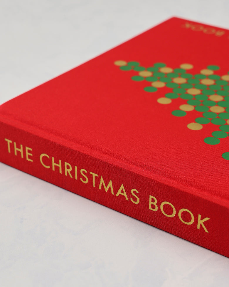 Close angled view of the red clothbound Christmas Book cover and spine, showing the gold title lettering and part of the tree design.
