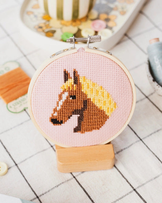 Horse head cross-stitch design in a wooden hoop on pink fabric, displayed on a small wooden stand.