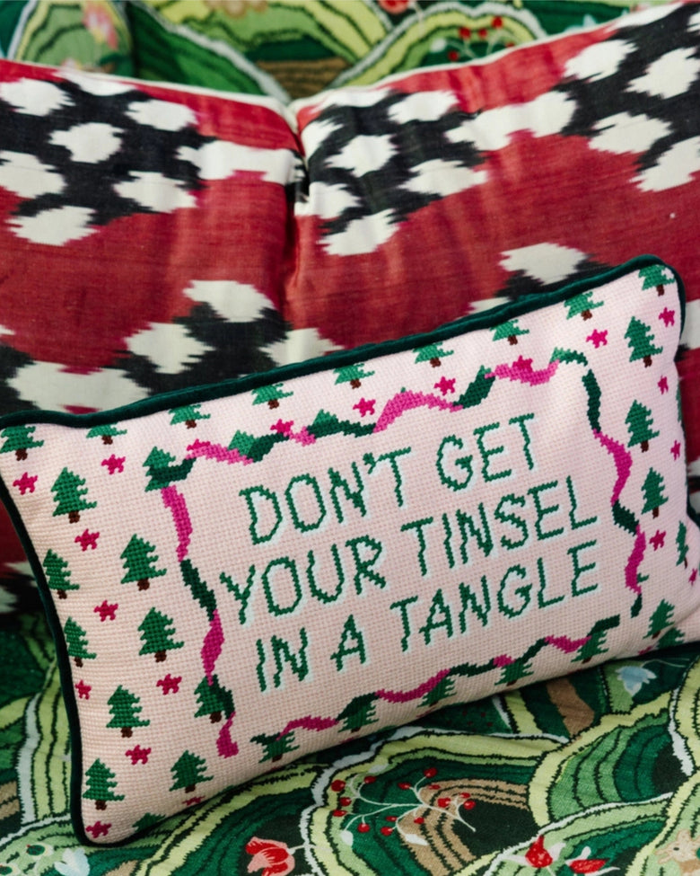 Pink needlepoint pillow reading “DON’T GET YOUR TINSEL IN A TANGLE,” shown against a holiday-patterned background.