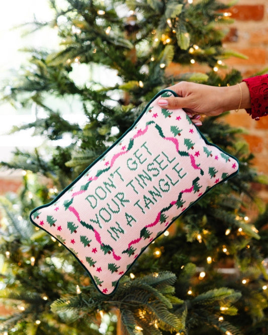 Pink needlepoint pillow reading “DON’T GET YOUR TINSEL IN A TANGLE,” held in front of a decorated Christmas tree.