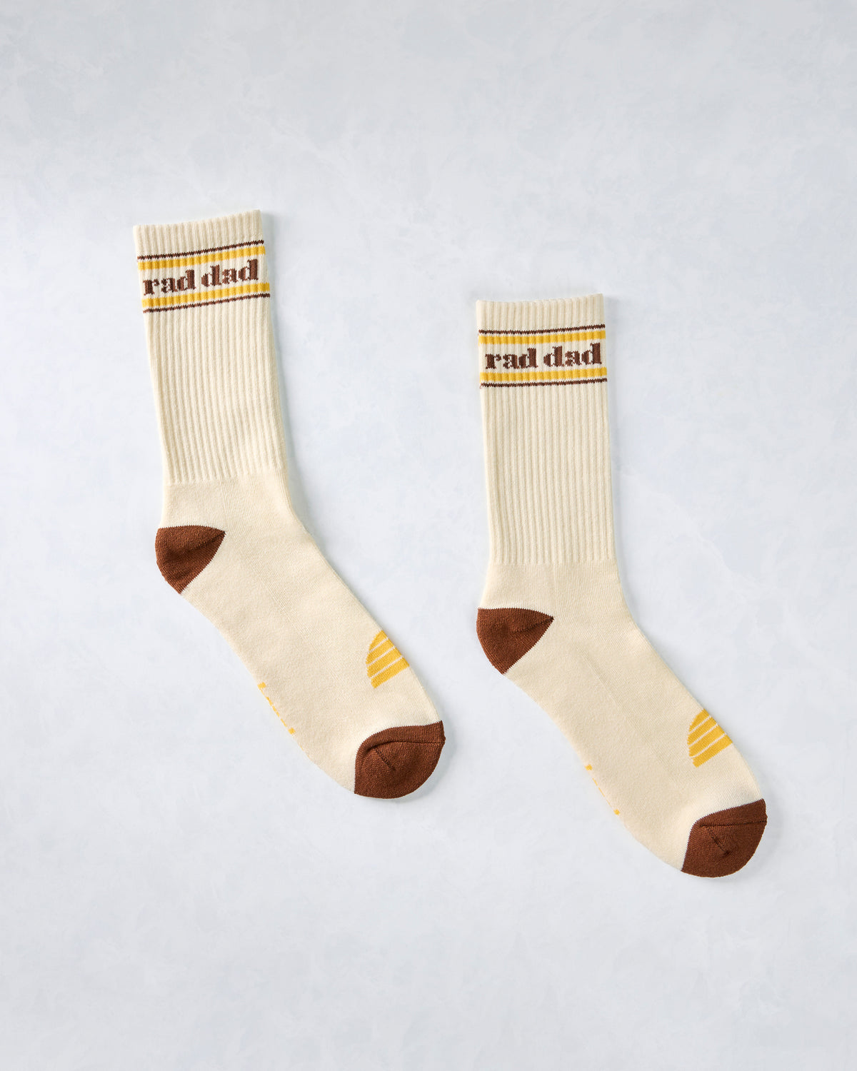 Pair of ivory crew socks displayed separately, with brown and yellow striped cuffs, “Rad Dad” lettering, brown heels and toes, and yellow accents.