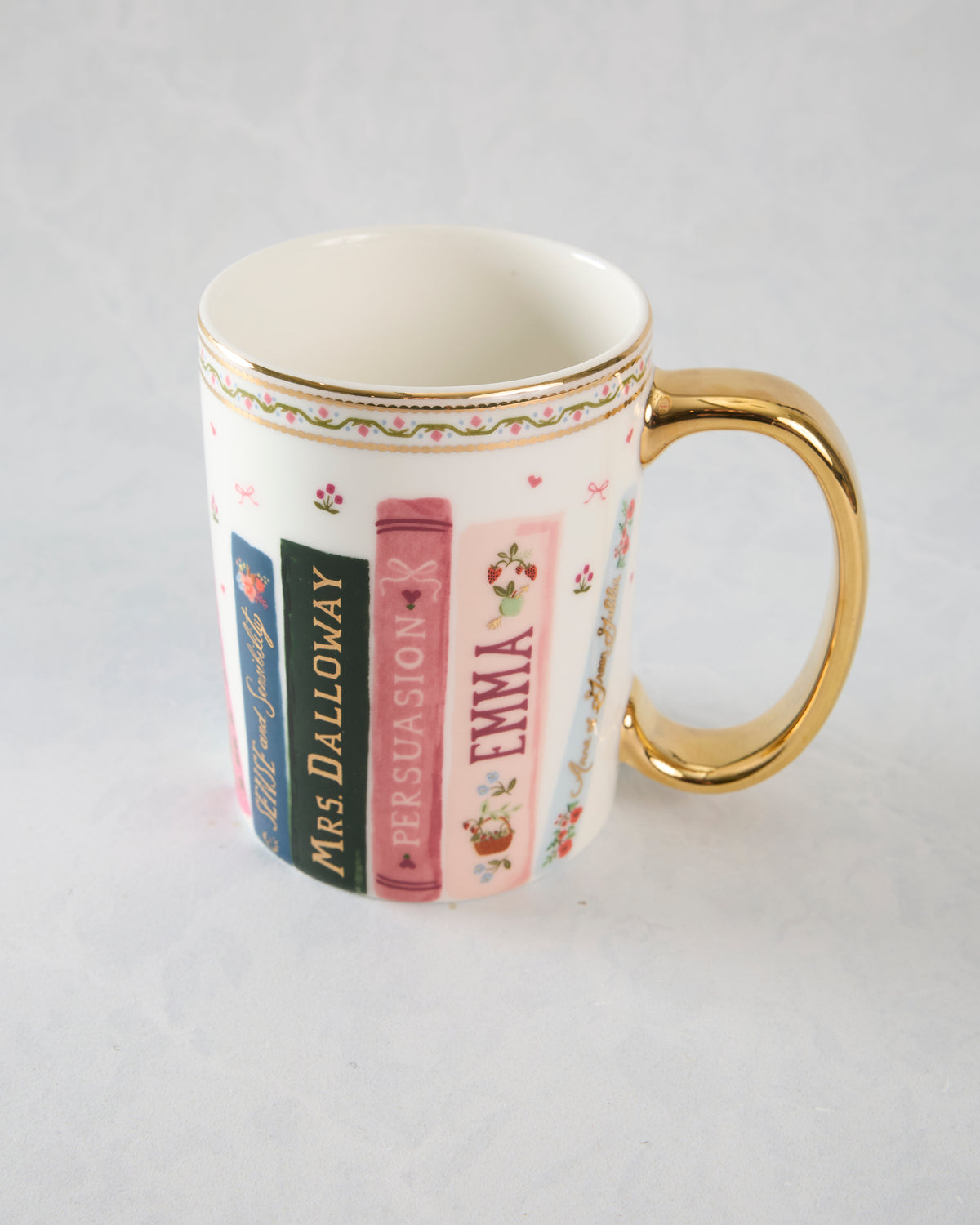 White porcelain mug with a gold handle and rim, decorated with illustrated book spines including Mrs. Dalloway, Persuasion, and Emma.