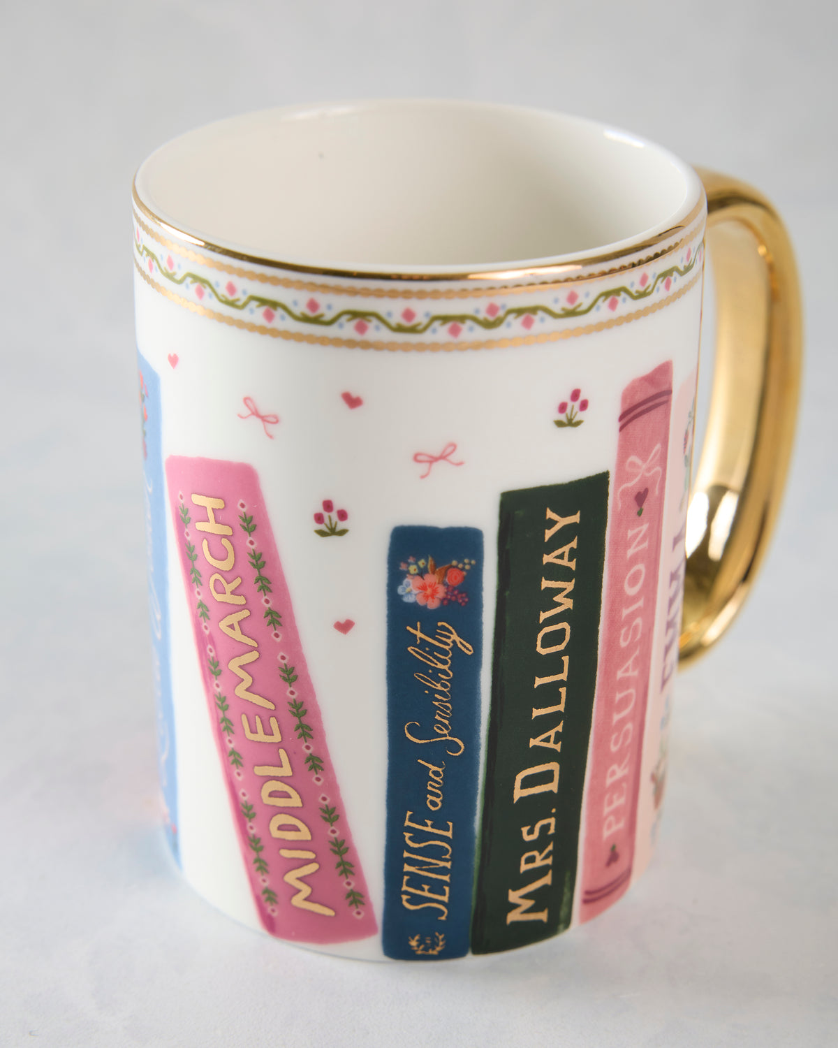 Porcelain mug with illustrated book spines, floral border, and metallic gold rim and handle.