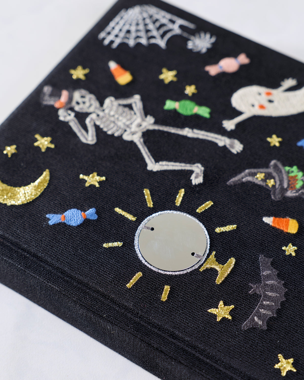 Angled close-up of the embroidered black journal cover, showing the skeleton, ghost, candies, stars, spiderweb, and round mirror-like detail.