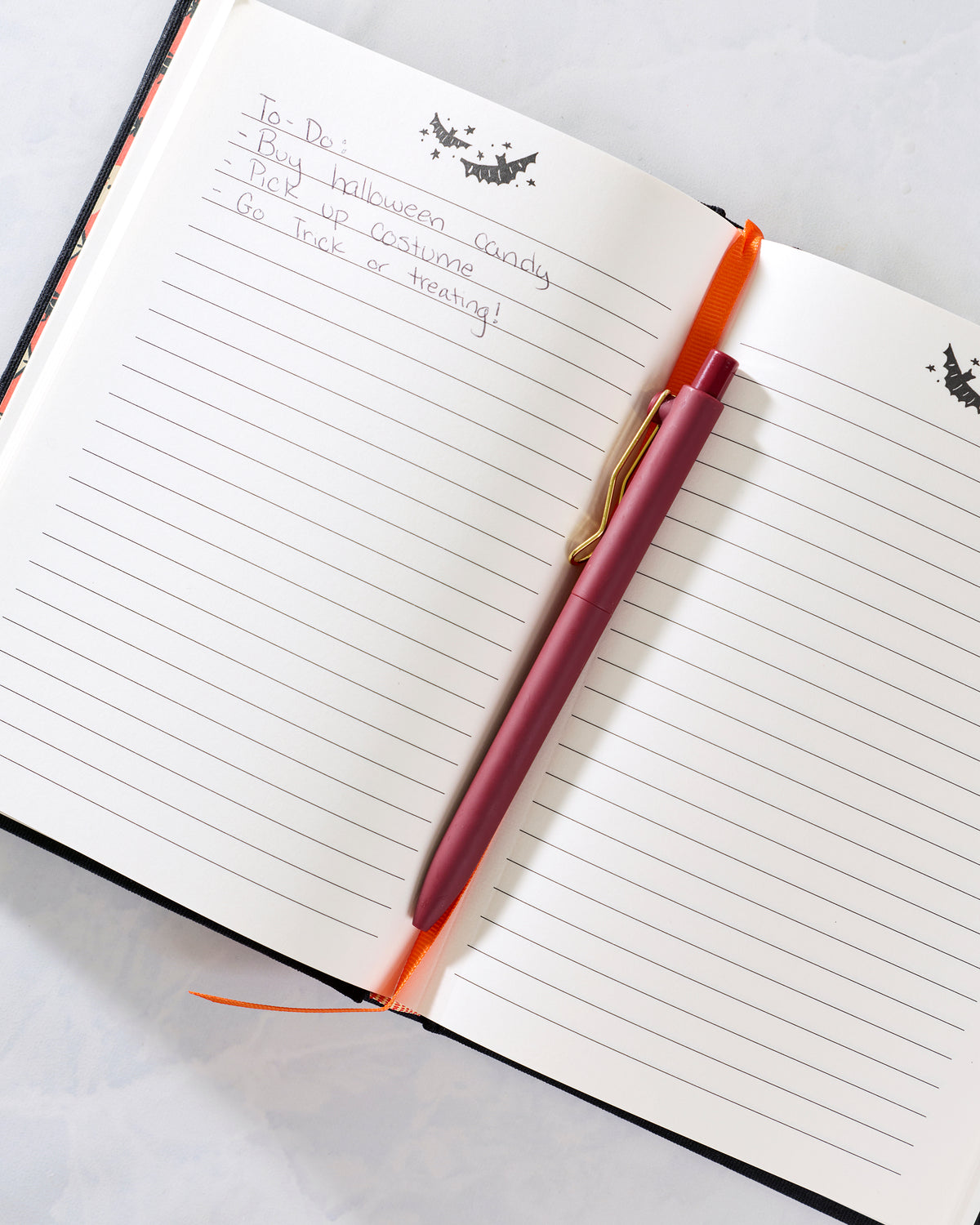 Open lined journal showing a handwritten Halloween to-do list, with a pink pen and orange ribbon bookmark.