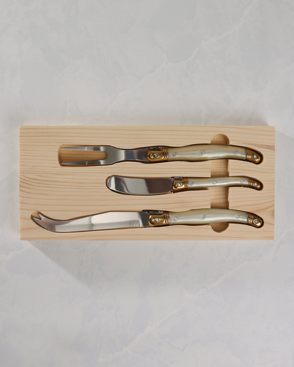 Three cheese knives with pearl-effect handles and gold-colored details arranged in a wooden tray.