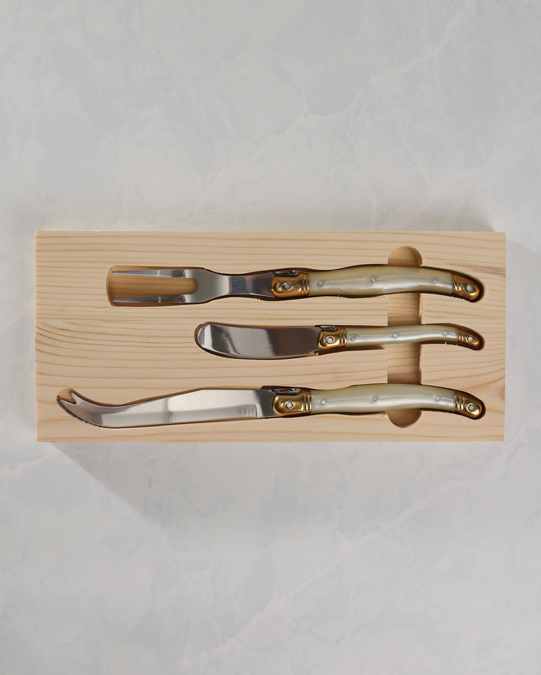 Three cheese knives with pearl-effect handles and gold-colored details arranged in a wooden tray.
