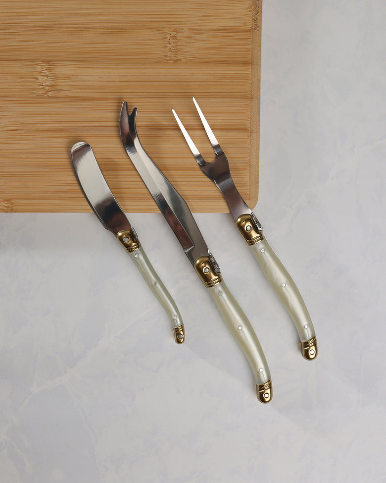 Three cheese knives with pearl-effect handles and gold-colored details displayed diagonally on a wooden board.