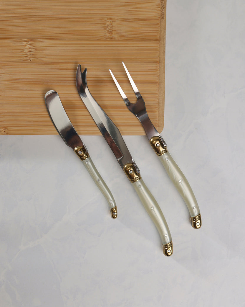 Three cheese knives with pearl-effect handles and gold-colored details displayed diagonally on a wooden board.