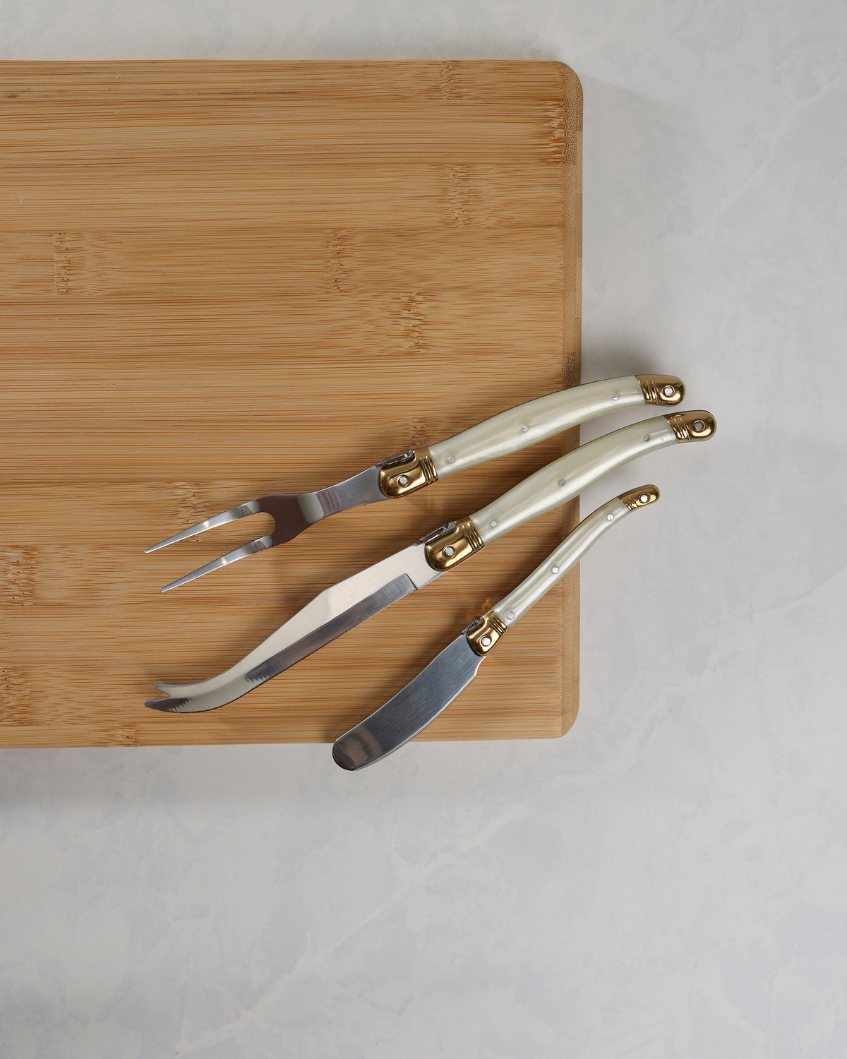 Three cheese knives with pearl-effect handles and gold-colored details arranged on a wooden board, showing their blades.