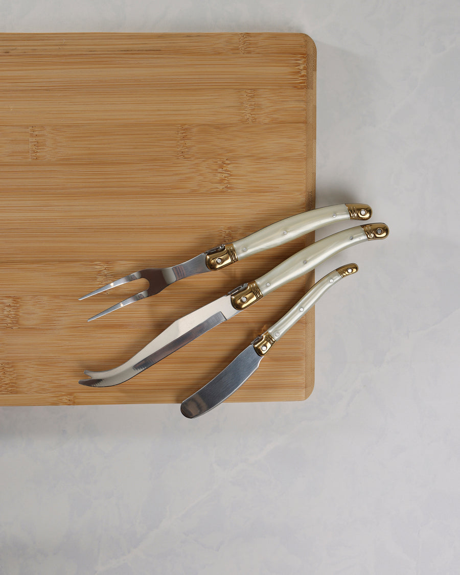Three cheese knives with pearl-effect handles and gold-colored details arranged on a wooden board, showing their blades.