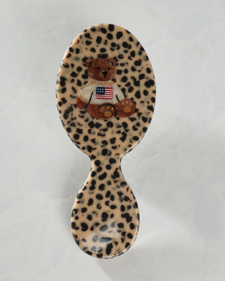 Back view of a leopard-pattern handbag hairbrush decorated with a teddy bear wearing a shirt with an American flag.