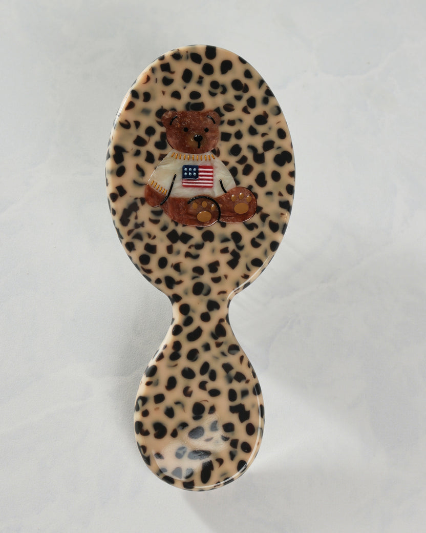 Back view of a leopard-pattern handbag hairbrush decorated with a teddy bear wearing a shirt with an American flag.