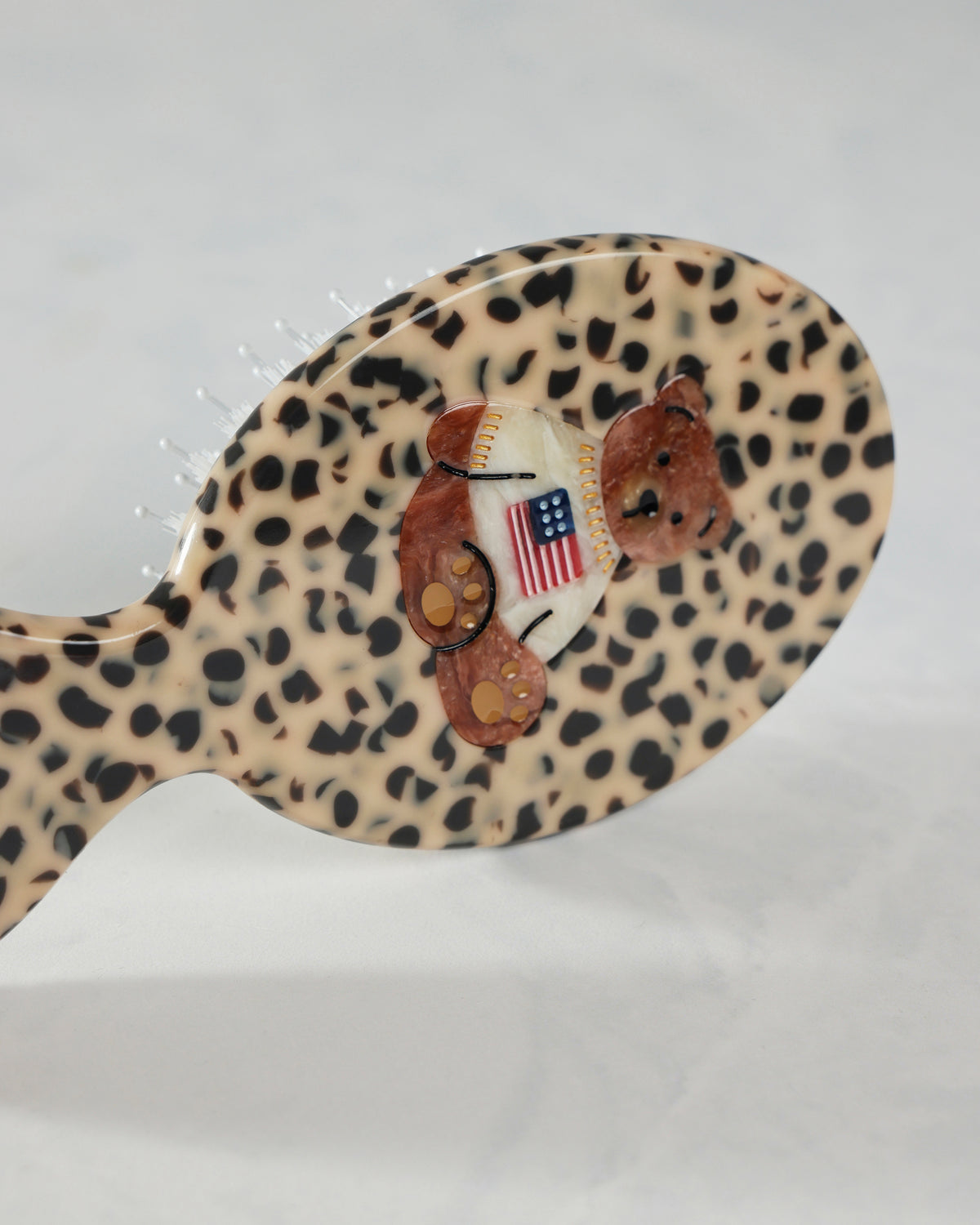 Angled close-up of the leopard-pattern hairbrush’s back, showing the raised teddy bear decoration and American flag shirt.