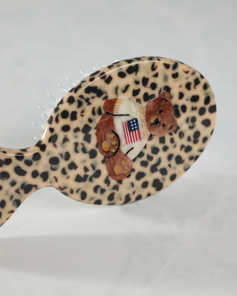 Angled close-up of the leopard-pattern hairbrush’s back, showing the raised teddy bear decoration and American flag shirt.