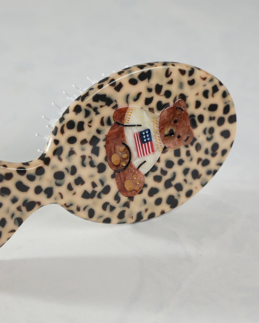 Angled close-up of the leopard-pattern hairbrush’s back, showing the raised teddy bear decoration and American flag shirt.