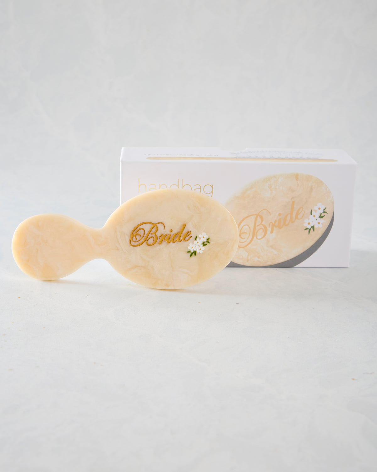 Cream handbag hairbrush with gold “Bride” lettering and small white flower decoration, displayed with its packaging.
