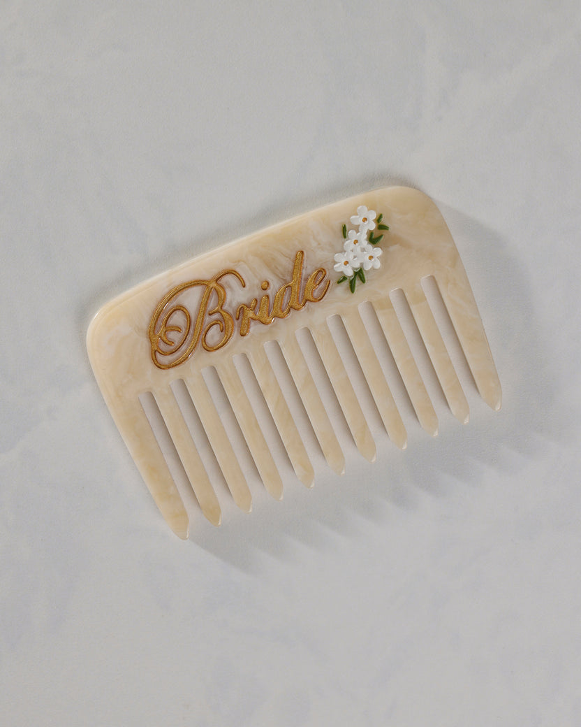 Cream pocket comb with wide rounded teeth, gold “Bride” lettering, and a small white floral decoration.