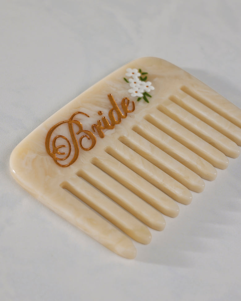 Close-up of a cream pocket comb showing its rounded teeth, gold “Bride” lettering, and white floral decoration.