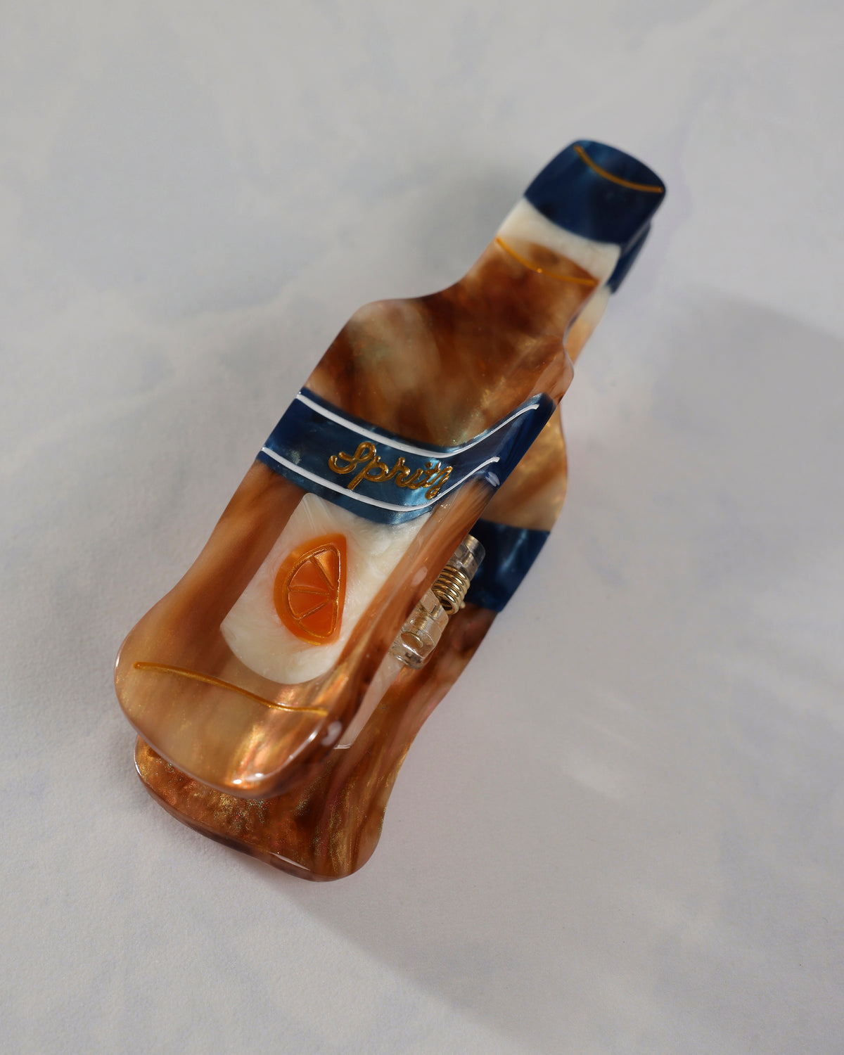 Angled underside view of a bottle-shaped citrus spritz claw hair clip, showing the spring hinge, citrus emblem, and navy, amber, and cream design.