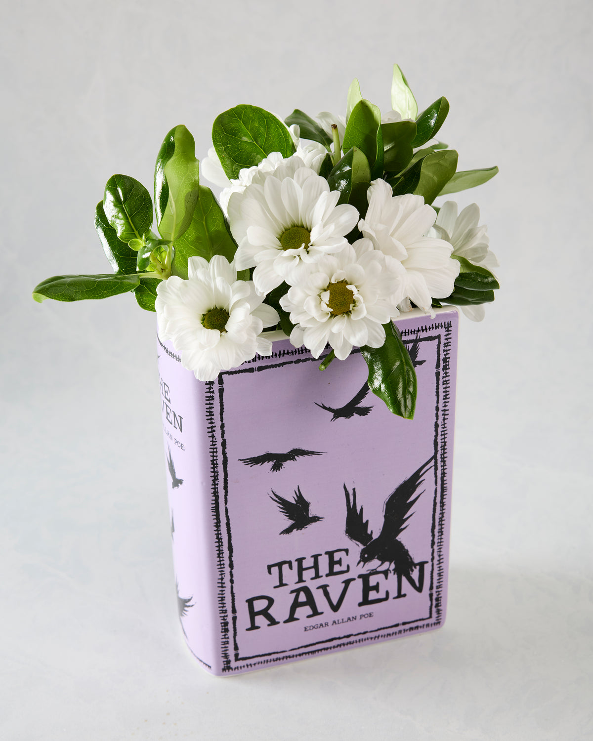 Purple book-shaped ceramic vase decorated with white flowers and green leaves, labeled The Raven.