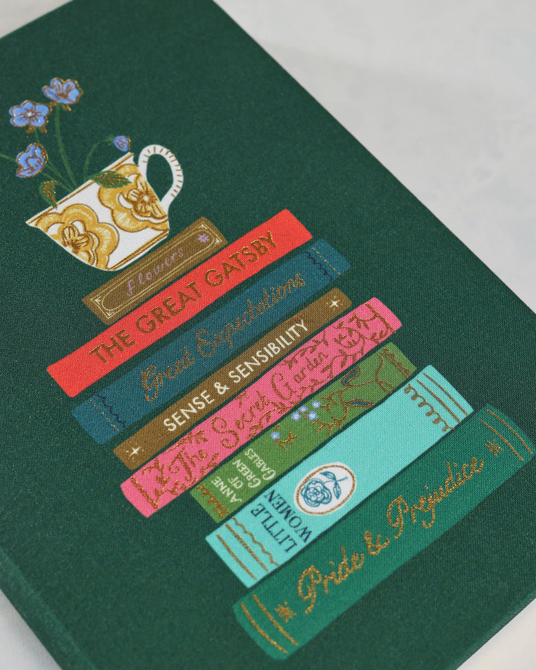 Angled close-up of the forest-green journal cover showing a floral teacup and stacked illustrated classic novels.