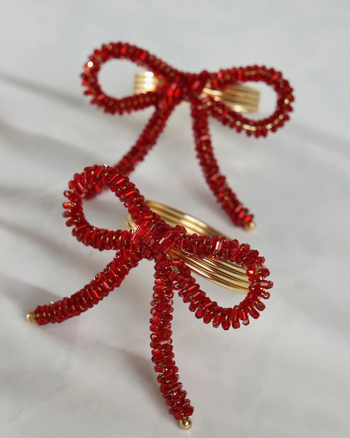 Two red beaded bow napkin rings with gold-tone metal centers, shown from an angled close-up view