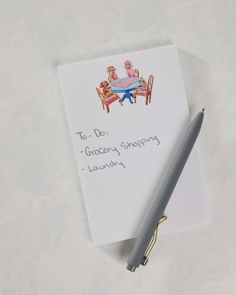 Notepad showing the dogs-around-a-table cover illustration and handwritten notes for grocery shopping and laundry, with a gray pen.