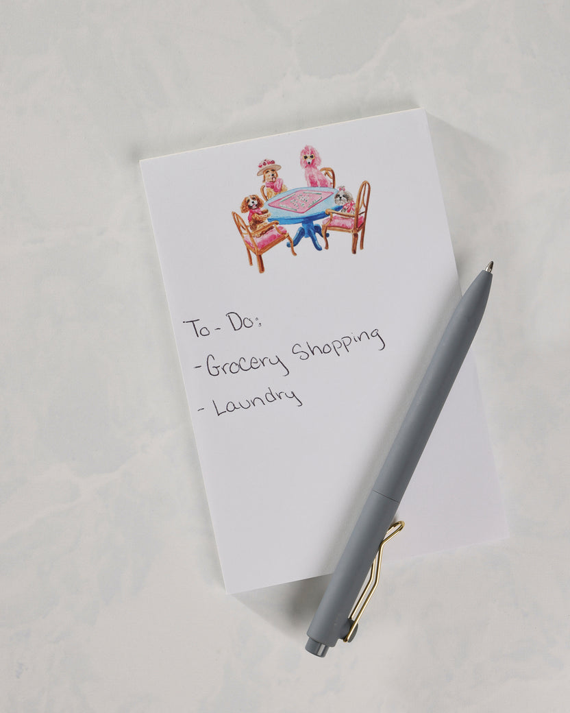Notepad showing the dogs-around-a-table cover illustration and handwritten notes for grocery shopping and laundry, with a gray pen.