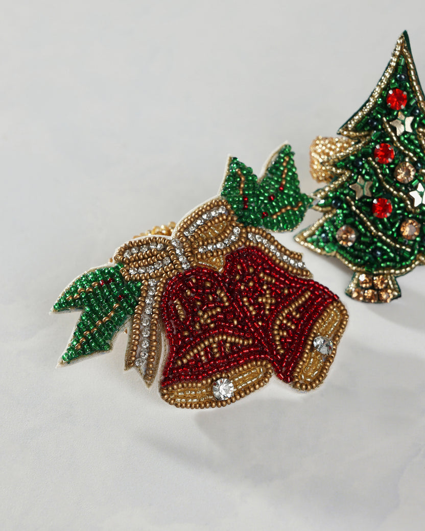 Close-up of a red bell-shaped napkin ring with green holly leaves, gold beadwork, and crystal accents beside a green Christmas tree design.
