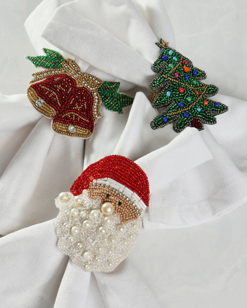 Three beaded holiday napkin rings—a red bell, green Christmas tree, and Santa—arranged on white cloth napkins.