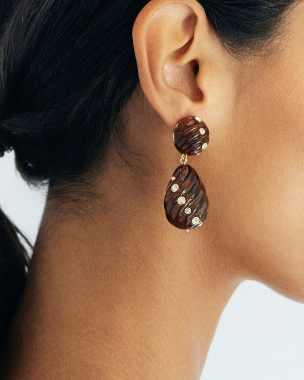 Close-up of an ear wearing a brown ridged tortoise drop earring with small clear stones.