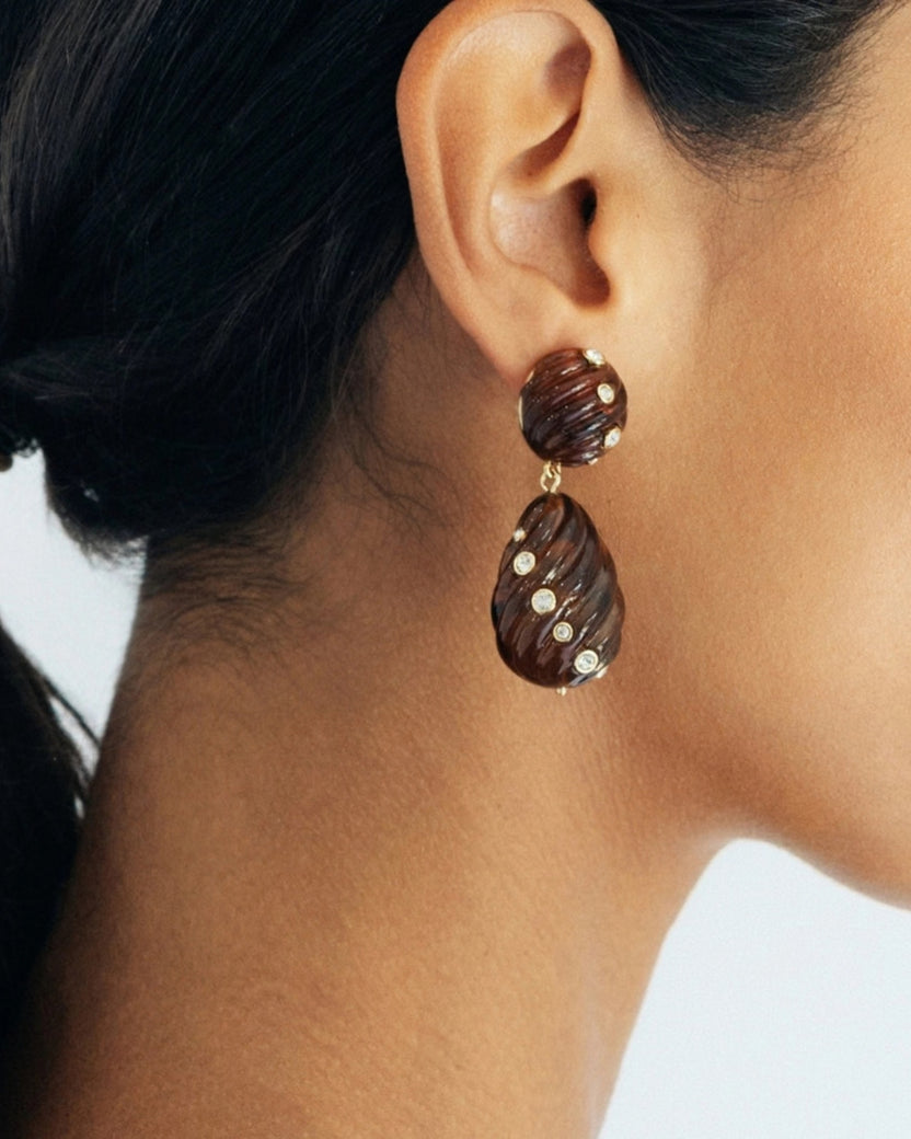 Close-up of an ear wearing a brown ridged tortoise drop earring with small clear stones.