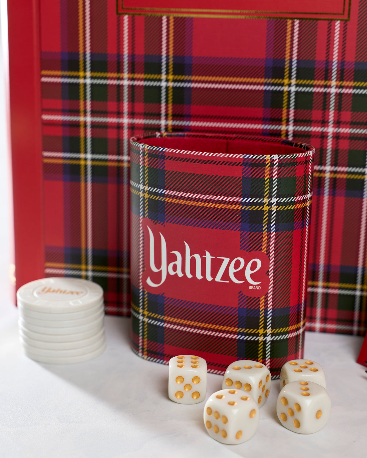 Close-up of the plaid Yahtzee dice cup with five white dice and stacked chips in front of the game case.