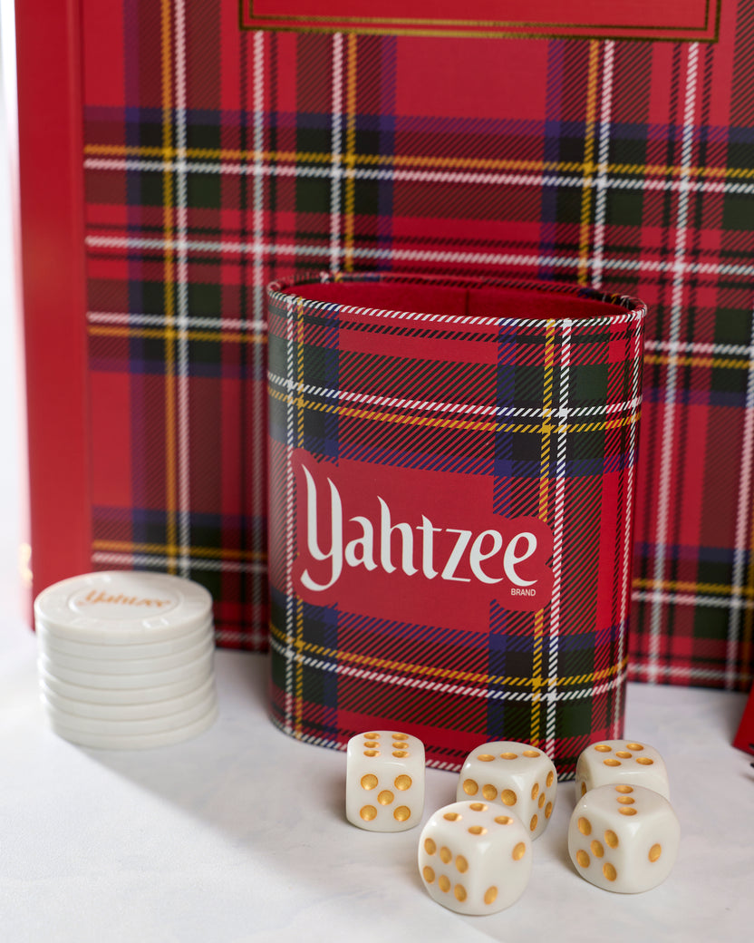 Close-up of the plaid Yahtzee dice cup with five white dice and stacked chips in front of the game case.