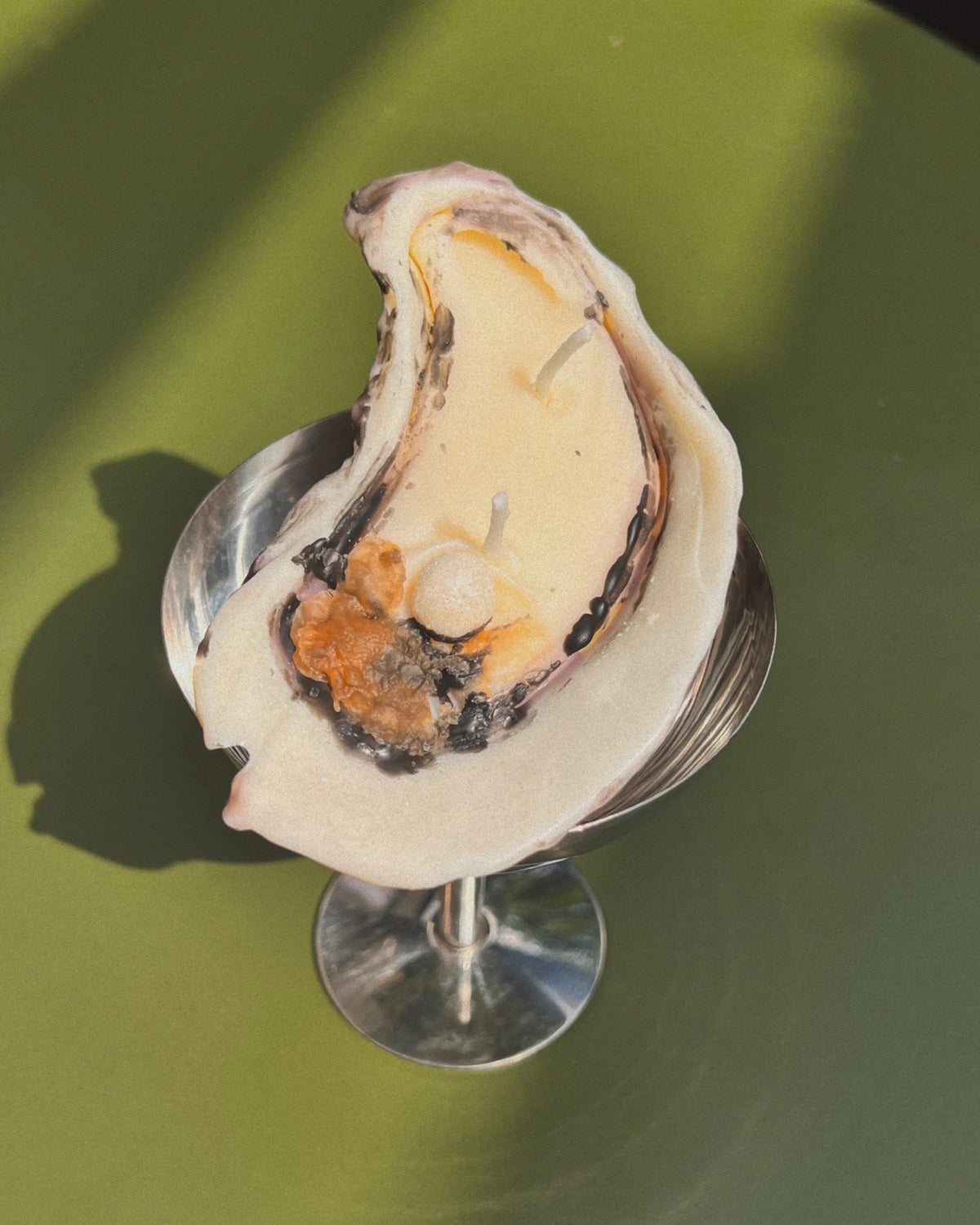 White oyster-shaped beeswax candle with a pearl detail in a coupe glass against a green background.
