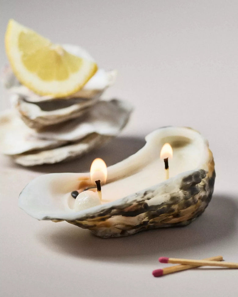 Lit oyster-shaped beeswax candle with two flames, oyster shells, a lemon wedge, and matches nearby.