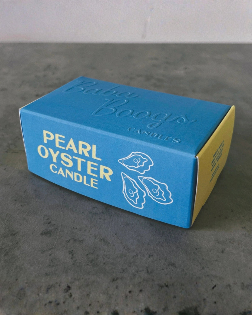 Blue and yellow packaging box for a Pearl Oyster Candle on a dark surface.