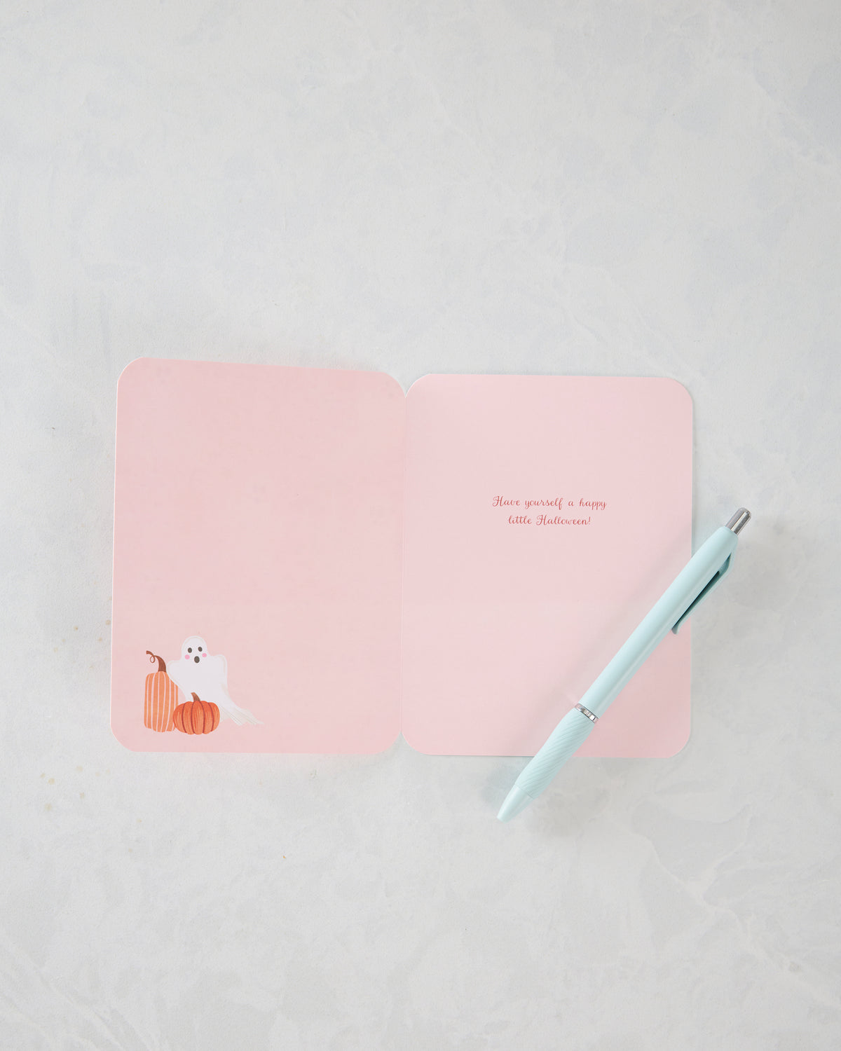 Open pink Halloween greeting card with a white ghost and orange pumpkins, shown beside a light blue pen; inside message reads, “Have yourself a happy little Halloween!”