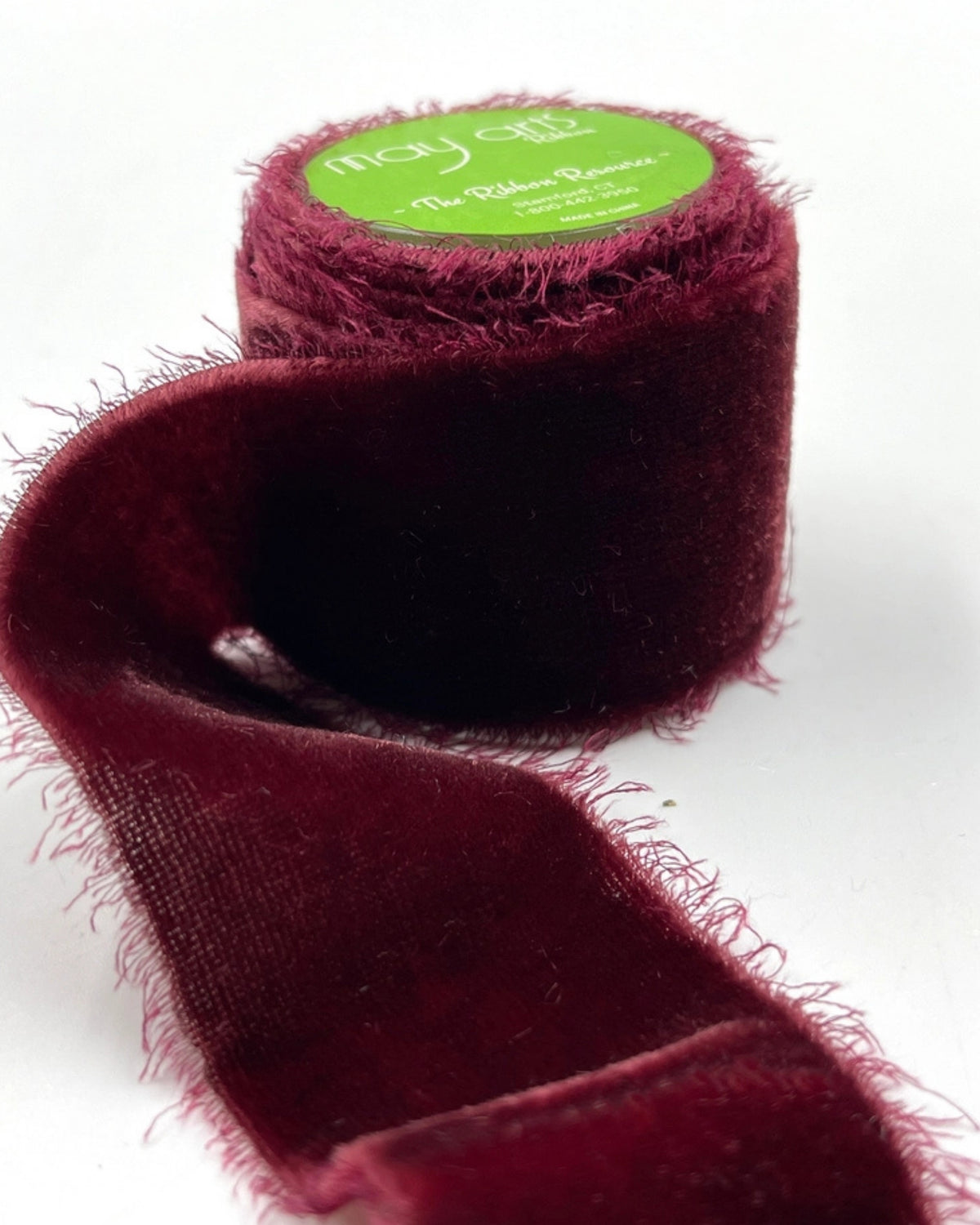 Roll of wine-colored silk velvet frayed ribbon with a loose length extending across a white background.